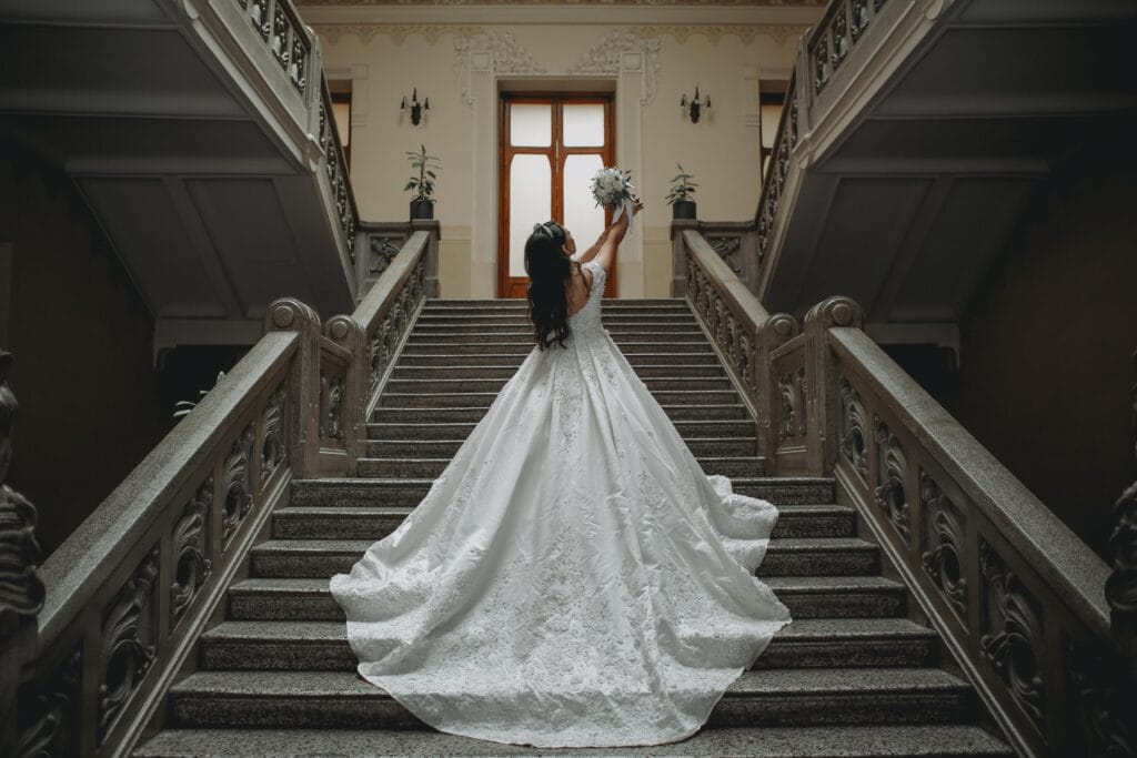 Bride wearing an elegant lace wedding gown with a dramatic train on a grand staircase, showcasing bridal gown inspiration for brides searching for beautiful wedding dresses.