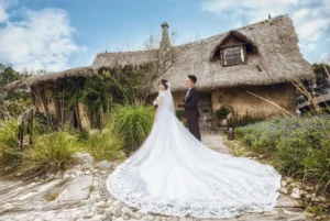 Alt text: Bride in a long, lace-trimmed white wedding gown with a dramatic train standing beside the groom in a black suit outside a rustic thatched-roof cottage, featured for “75 Jaw-Dropping Wedding Dresses Every Bride-to-Be Should See Before Saying ‘I Do’.”