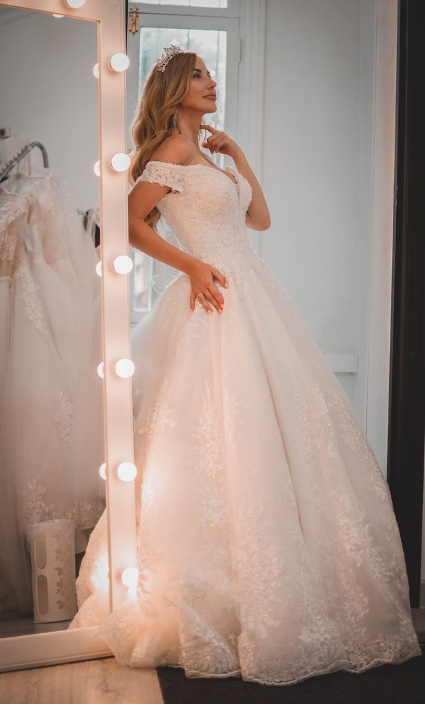 Bride trying on a lace wedding dress while planning a wedding dress budget, with a full bridal gown and tiara in a fitting room.