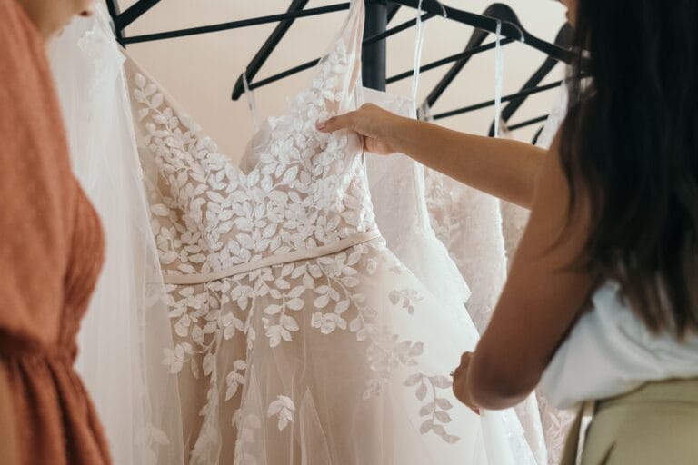 Bride shopping for a wedding dress in a bridal boutique, looking at a floral lace wedding gown among other dresses.