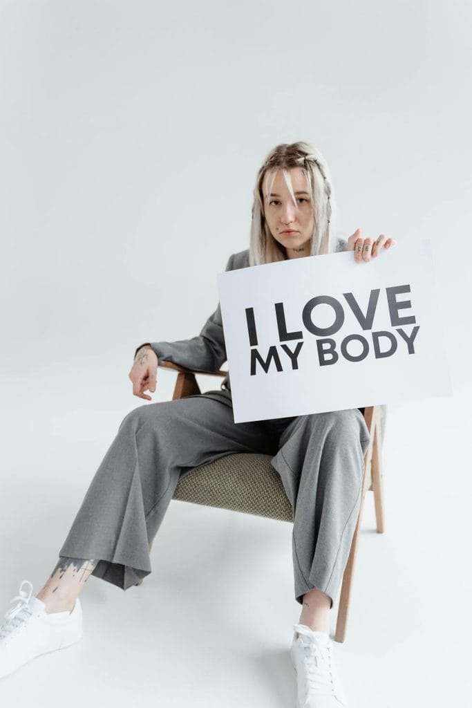 Woman holding an I Love My Body sign, emphasizing body confidence for petite women while encouraging them to feel confident as they learn how to dress to look taller.