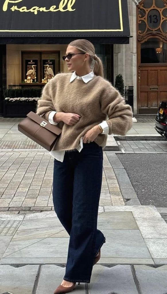 Woman wearing a gray crewneck sweatshirt with a blue collared shirt, dark straight-leg jeans, a brown belt, and black shoes for sweater and jeans outfit ideas.