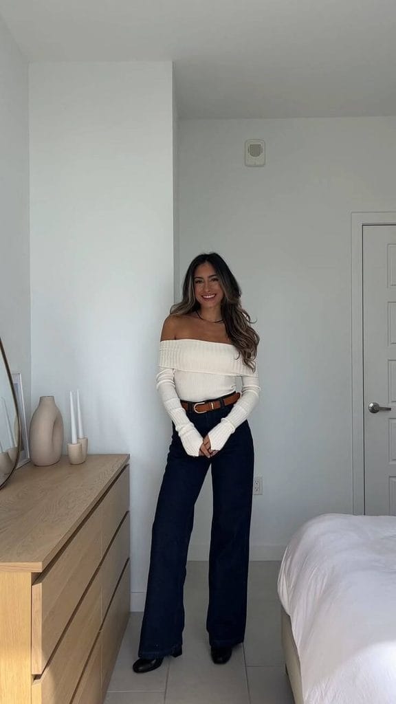 Woman wearing a white off-the-shoulder sweater with dark wide-leg jeans and black shoes for sweater and jeans outfit ideas.