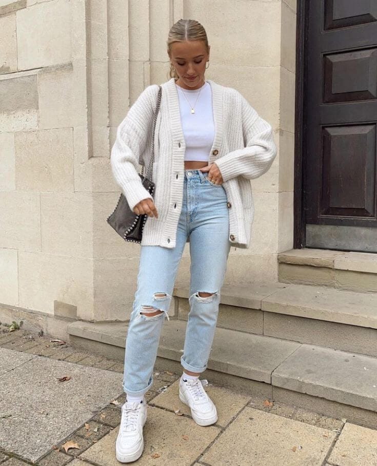 Woman wearing an oversized white cardigan with a white top, distressed jeans, and white sneakers for cardigan and jeans outfit ideas.