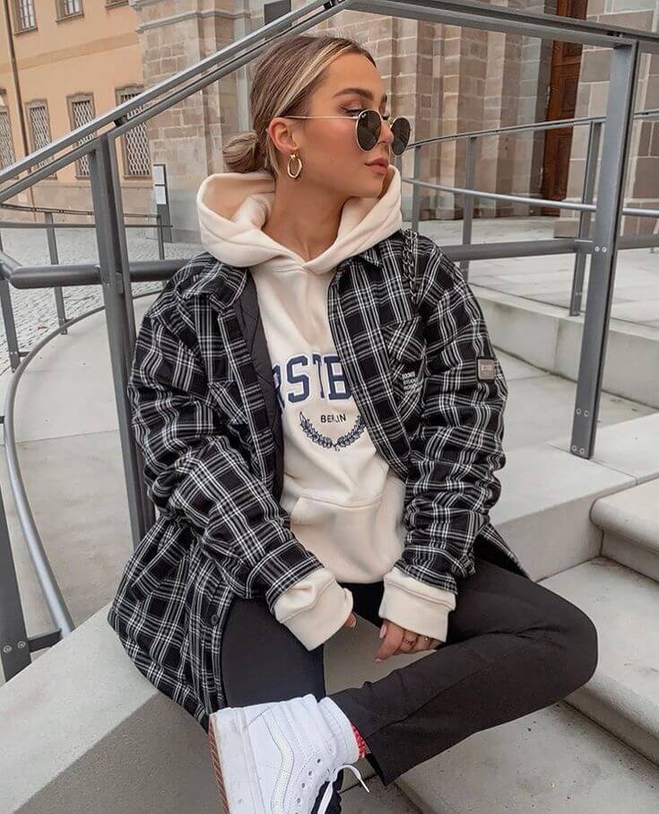 Woman wearing a black plaid shacket over a cream hoodie with black leggings and white sneakers showing shacket outfit ideas.