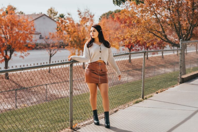 Woman wearing a cream button-front sweater, a brown leather mini skirt, and black ankle boots against a backdrop of orange autumn trees, representing 20 Thanksgiving dinner outfit ideas for women.