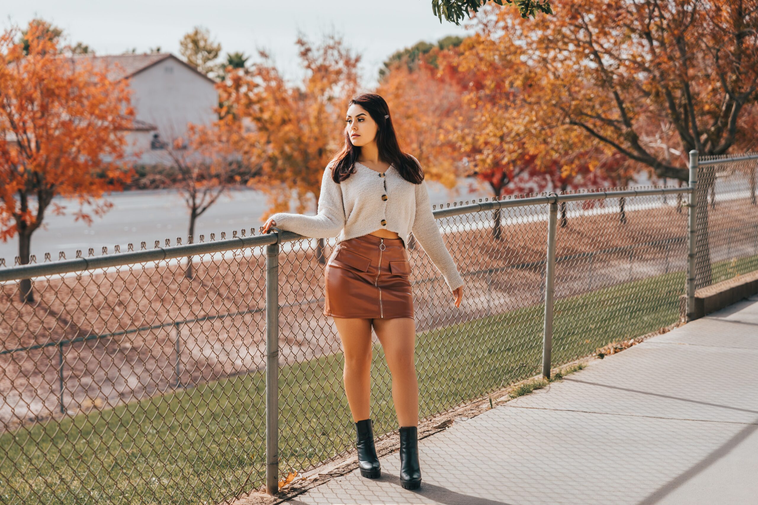 Woman wearing a cream button-front sweater, a brown leather mini skirt, and black ankle boots against a backdrop of orange autumn trees, representing 20 Thanksgiving dinner outfit ideas for women.