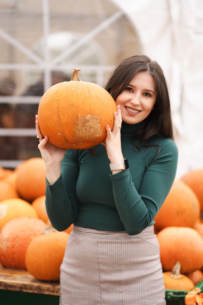 Woman wearing a dark green turtleneck and a light plaid skirt while holding an orange pumpkin, illustrating the best colors to wear to a pumpkin patch.