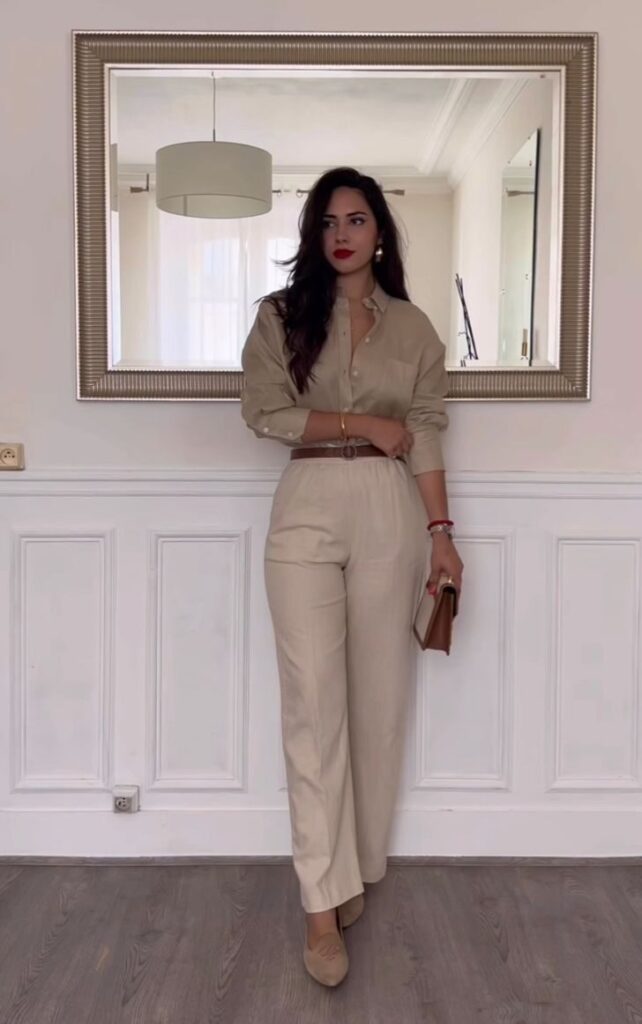Button-down shirt and tailored pants outfit for Thanksgiving dinner featuring a woman wearing a beige button-down shirt with matching tailored pants, a brown belt, beige flats, and a brown clutch.
