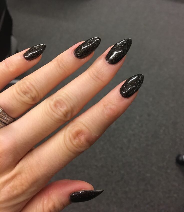 Glossy black almond-shaped nails with fine glitter scattered across the surface, creating classic black glitter nails for Halloween and a sparkling fall nail idea.