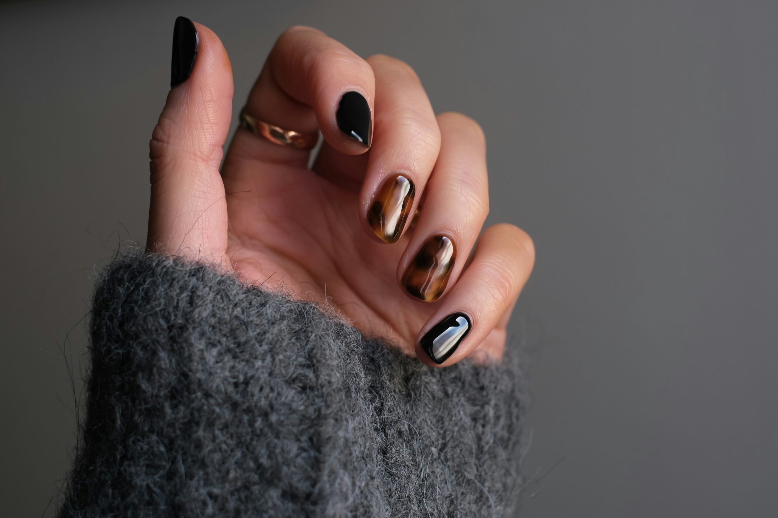 A hand featuring glossy black nails with brown tortoiseshell accent nails and a cozy gray knitted sweater, representing fall nail ideas for early fall, autumn, Halloween, and Thanksgiving, with seasonal manicure inspiration for the fall season.