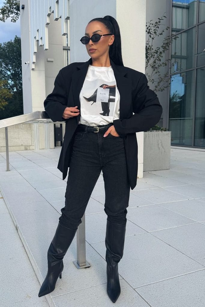 Woman wearing a white graphic Halloween T-shirt with black straight-leg jeans, a long black blazer, and black knee-high boots, showing how to style a Halloween T-shirt with an all-black outfit.