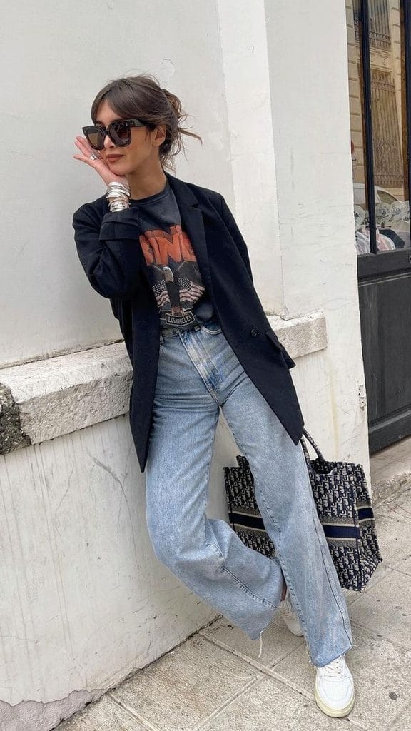 A woman wearing a black Halloween graphic T-shirt with a black blazer, relaxed-fit jeans, white sneakers, sunglasses, and a tote bag, showing how to style a Halloween T-shirt with a blazer and jeans for a casual outfit look.