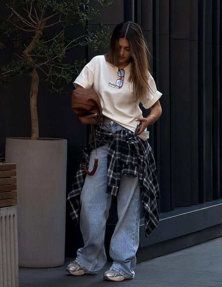 Woman wearing a white graphic T-shirt with relaxed boyfriend jeans and an oversized plaid flannel tied around her waist, showing how to style a Halloween T-shirt with boyfriend jeans and a plaid flannel for a relaxed 90s-inspired fall look.