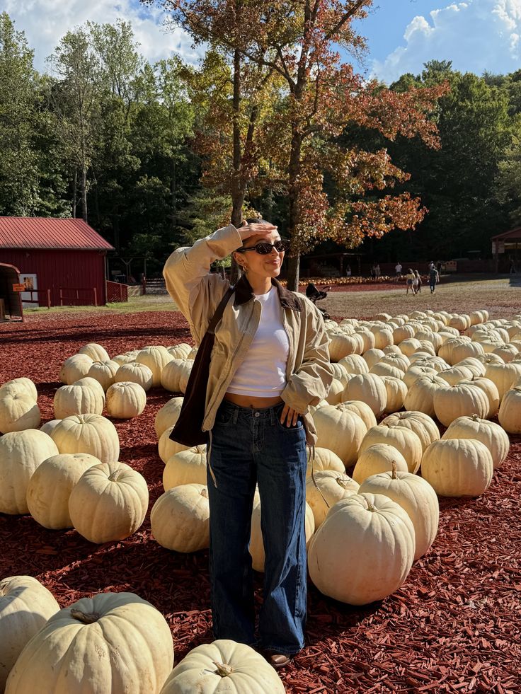Woman wearing a beige jacket over a white cropped top with dark-wash wide-leg jeans and sunglasses at a pumpkin patch, showcasing a layered shirt outfit for a pumpkin patch.