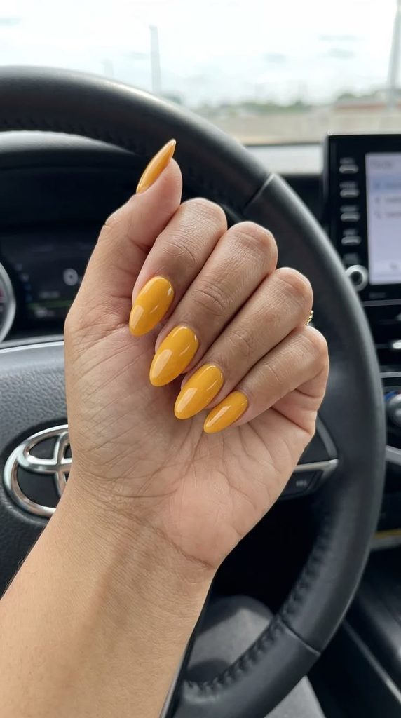 A hand with long almond-shaped nails painted in a solid marigold yellow shade with a glossy finish, paired with gold rings and a mustard-colored ribbed sweater in warm autumn sunlight, showcasing marigold yellow nails for fall nail ideas.