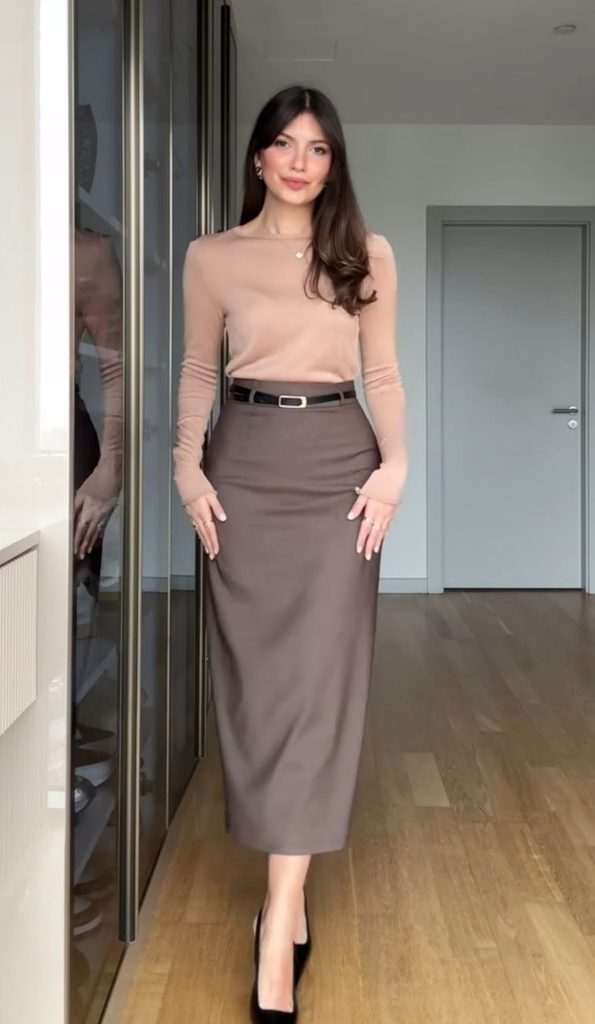 Woman wearing a beige fitted long-sleeve top tucked into a taupe midi skirt with a black belt and black heels, showcasing a midi skirt and fitted top outfit for Thanksgiving dinner.