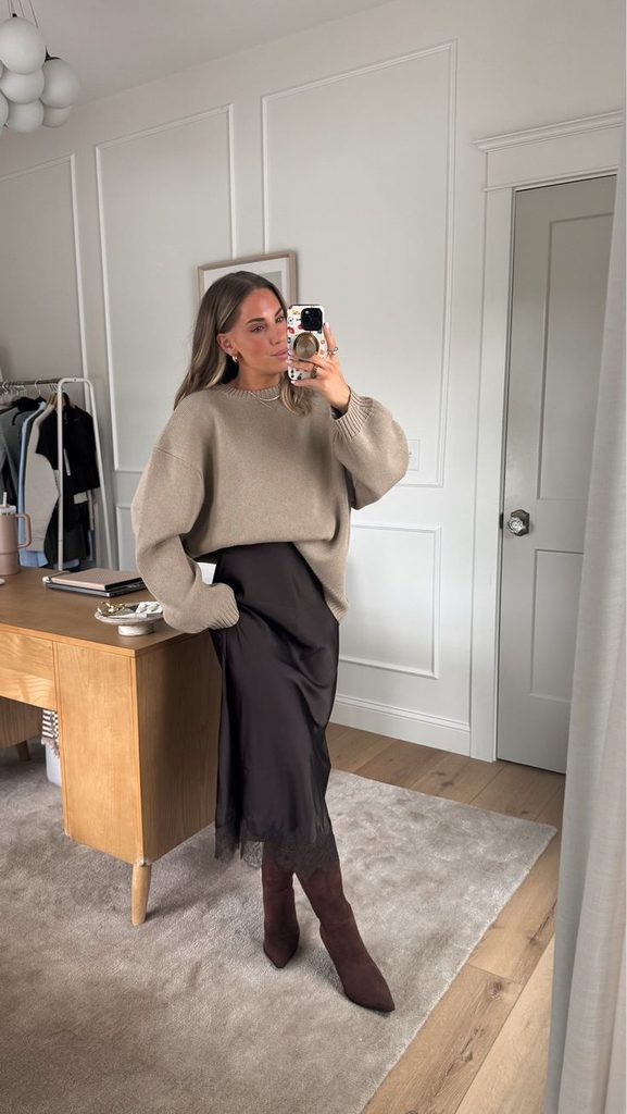Midi slip dress and sweater outfit for Thanksgiving dinner featuring a woman wearing an oversized beige sweater over a black satin midi slip dress, paired with burgundy knee-high boots.