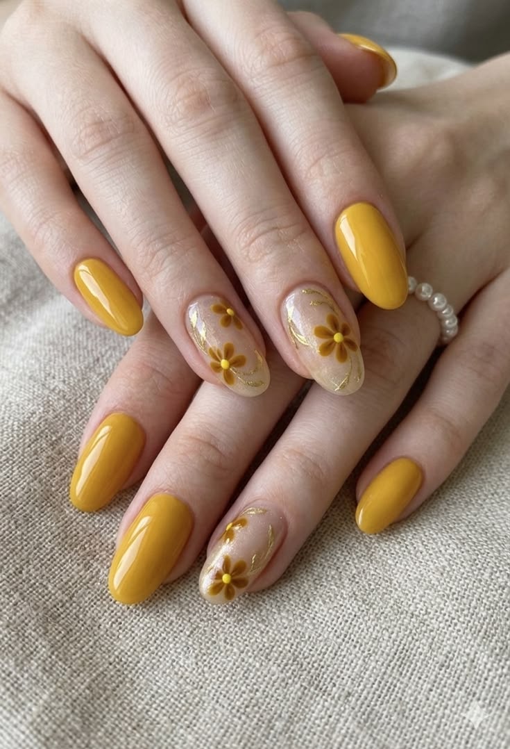 A hand with glossy mustard-yellow floral fall nails, featuring solid mustard-yellow polish on most nails and delicate brown and yellow daisy designs with fine gold accents on sheer nude accent nails, creating an elegant autumn manicure.