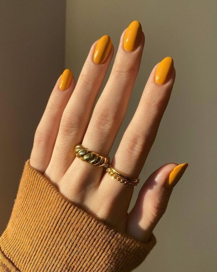 A hand with long almond-shaped nails painted in a solid mustard yellow shade with a glossy finish, paired with gold rings and a mustard-colored ribbed sweater in warm autumn sunlight, showing mustard yellow nails for fall nail ideas.