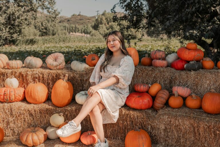 Woman posing in a patterned dress and white sneakers among colorful pumpkins at a pumpkin patch, capturing the inspiration behind pumpkin patch outfit ideas for women.