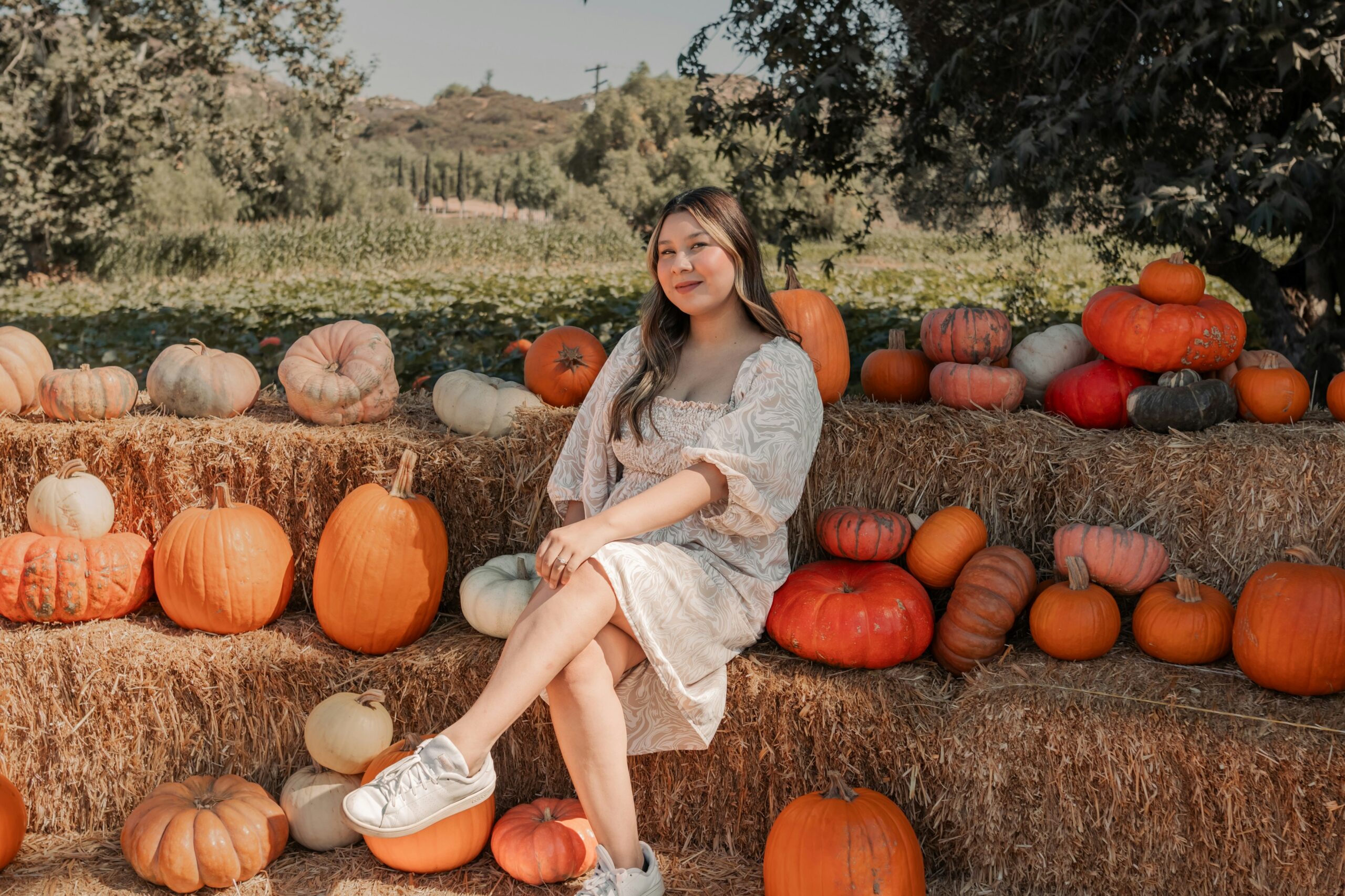 Woman posing in a patterned dress and white sneakers among colorful pumpkins at a pumpkin patch, capturing the inspiration behind pumpkin patch outfit ideas for women.