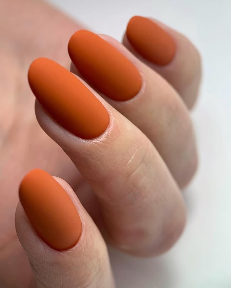 Close-up of matte pumpkin-orange polish on medium-length, rounded almond-shaped nails, showcasing a simple fall manicure in a warm burnt-orange shade. These pumpkin spice nails are a seasonal fall nail idea for September and October.
