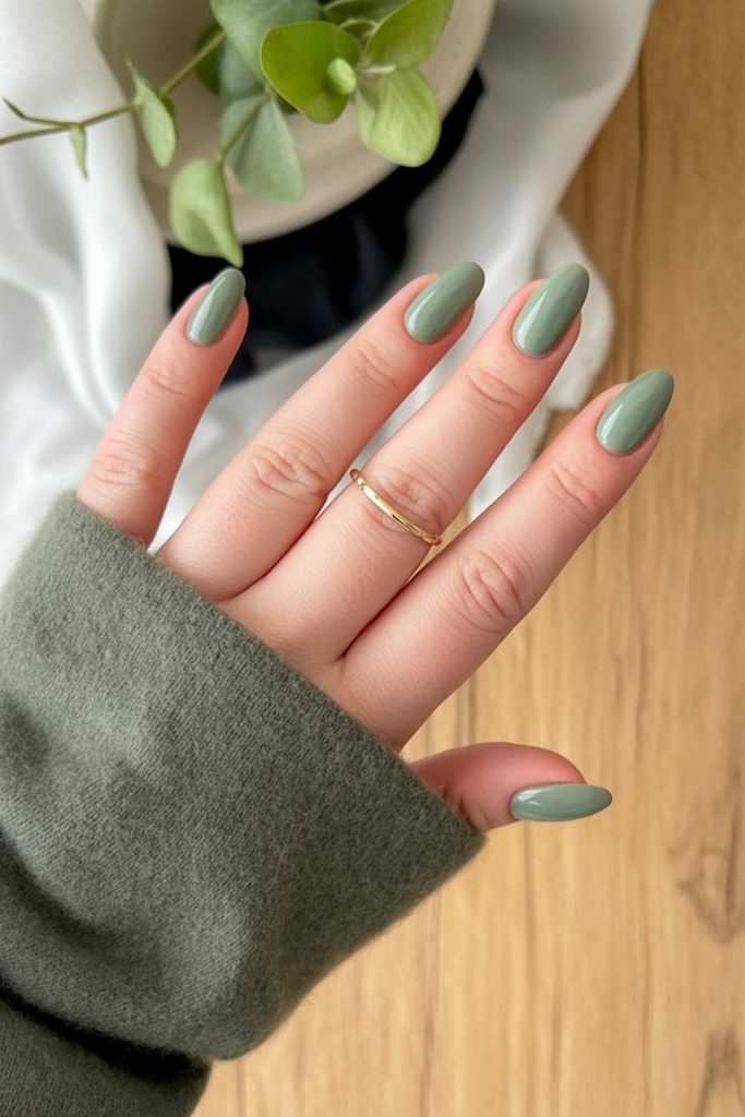 Close-up of a hand with glossy sage-green almond-shaped nails, a thin gold ring, and a cozy green knit sweater, showcasing sage green nails for fall nail ideas.
