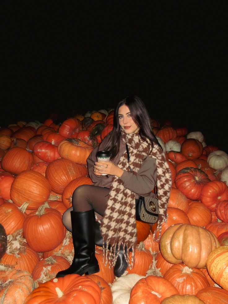 Woman wearing a brown outfit with a brown-and-cream patterned scarf, black knee-high boots, and a crossbody bag while posing among pumpkins at a pumpkin patch.