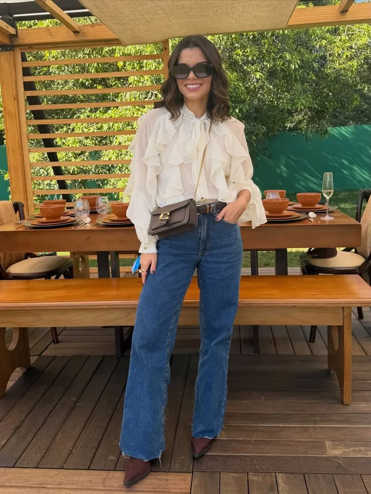 Woman wearing dark blue straight-leg jeans, a white ruffle blouse, brown pointed-toe ankle boots, and a brown crossbody bag for a dressy casual Thanksgiving dinner outfit.