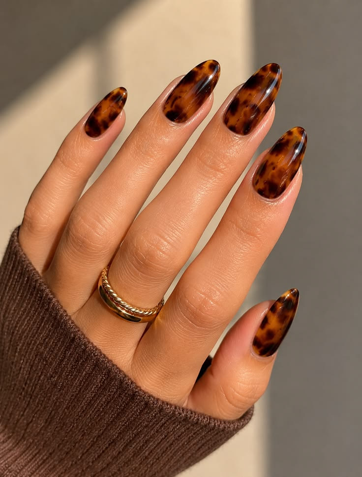 Close-up of long almond-shaped nails featuring a glossy tortoiseshell pattern in amber, caramel, and dark brown, paired with gold rings and a brown knit sweater, showcasing tortoiseshell nails for fall and fall nail ideas.