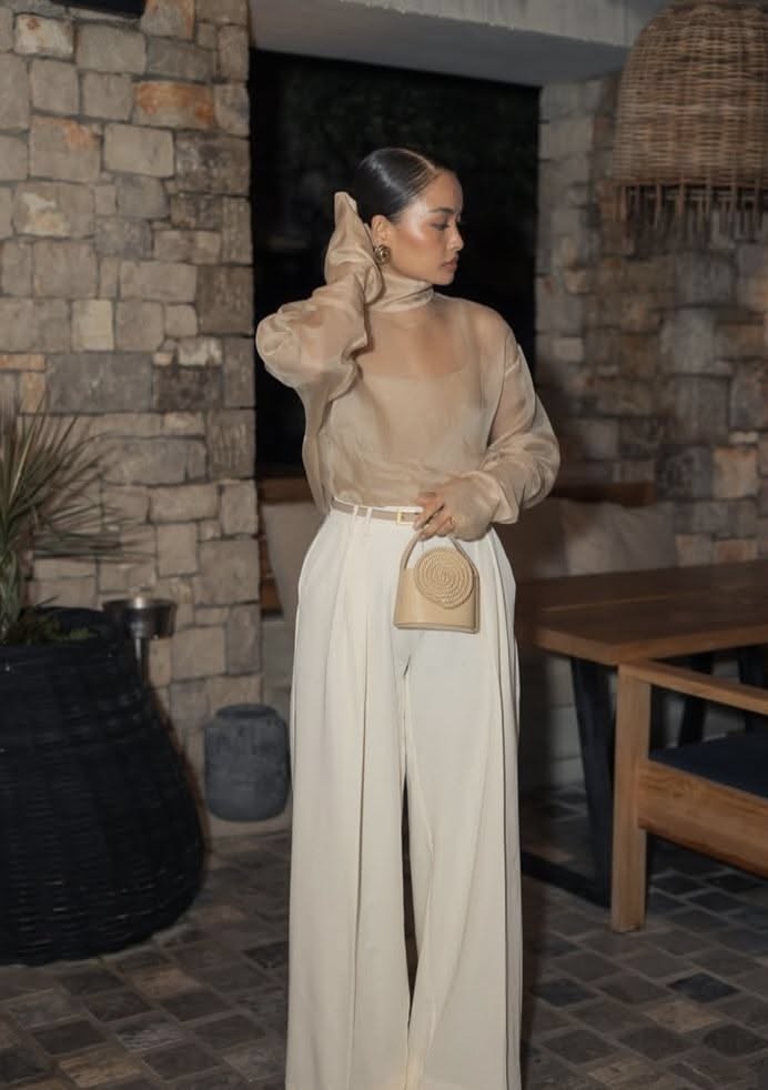 Woman wearing a sheer beige long-sleeve blouse tucked into white wide-leg pants with a matching belt and a beige handbag, showcasing a dressy wide-leg pants outfit for Thanksgiving dinner.